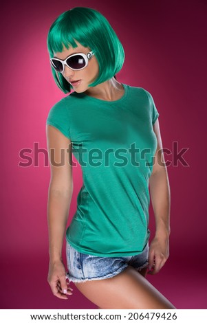 Cute slender young woman with psychedelic green hair in a matching green top, trendy sunglasses and skimpy denim shorts looking down to the side