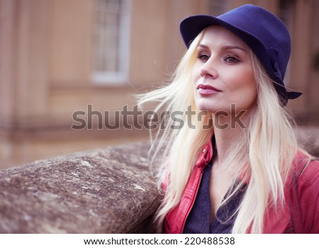 Stylish young blond woman waiting outdoors with her long hair blowing in the breeze leaning against a stone wall looking off to the left of the frame