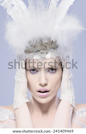 Fine art cool toned portrait of a gorgeous winter maiden wearing a feathery white headdress with her hands in lacy gloves held to her head, closeup head and shoulders