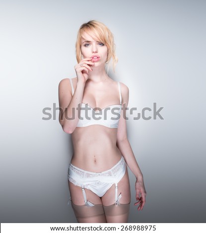 Sexy Pouting Young Blond Woman Wearing White Bra, Panties, Garter Belt and Stockings Standing in Studio with Grey Background