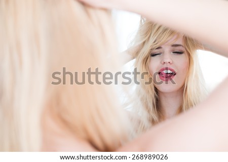 Young Blond Woman with Hands in Hair Licking Lips and Looking into Mirror, as seen from Behind