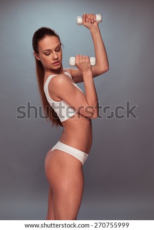 Portrait of a Sexy Young Woman, Wearing White Underwear, Raising Arms with Dumbbells on a Gray Background.