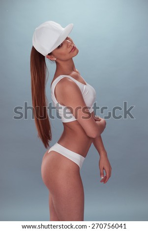 Portrait of a Sexy Long-Haired Woman, Wearing White Underwear with Cap, Crossing her Arm Over her Stomach While Looking at the Camera. Isolated on a Gray Background. Captured in Side View.