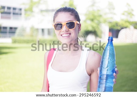 Close up Pretty Young Woman in a Summer Outfit at the Park, Holding a Plastic Bottle of Water and Smiling at the Camera.