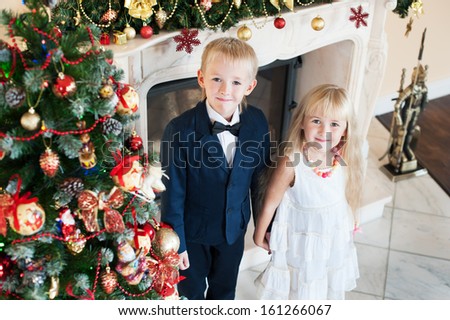 Happy brother sister holding hands near the Christmas tree and fireplace. children in evening dresses celebrate New year