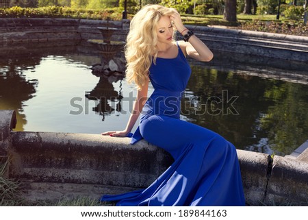 beautiful woman with blond hair in elegant blue dress sitting beside a lake at summer park