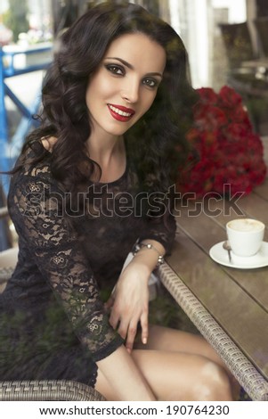 beautiful smiling woman with dark hair in black lace dress sitting in city cafe with cup of coffee and bouquet of red roses on background