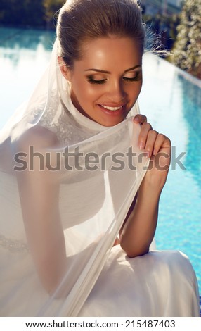 fashion photo of beautiful smiling bride with blond hair in elegant wedding dress posing beside a swimming pool
