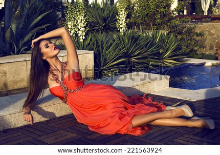 fashion outdoor photo of beautiful girl with long dark hair in luxurious red dress relaxing at villa