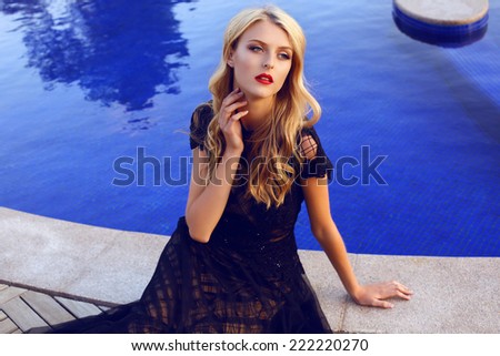 fashion outdoor photo of beautiful girl with blond curly hair in elegant lace dress posing beside a swimming pool