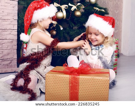 positive photo of cute beautiful little girls in Santa\'s hat playing beside a decorated Christmas tree and presents