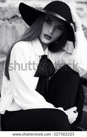 black and white fashion outdoor photo of beautiful ladylike woman with dark hair wearing elegant blouse and black hat