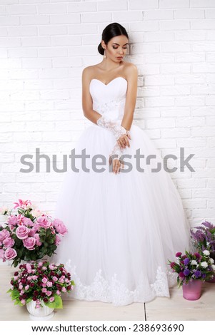 fashion studio photo of beautiful bride with dark hair in elegant wedding dress posing among bouquets of flowers