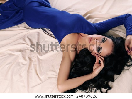 fashion photo of sexy glamour woman with dark hair wearing elegant blue dress, lying on bed in bedroom
