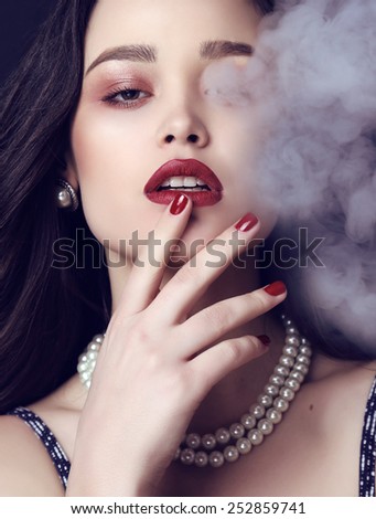 fashion studio photo of beautiful sensual woman with dark hair and bright makeup,with pearls necklace posing in cigarette\'s smoke