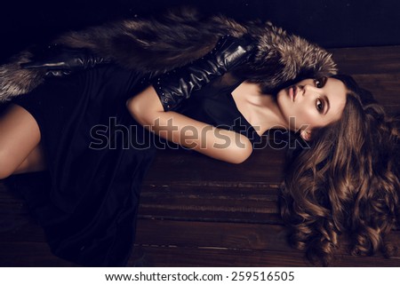 fashion photo of sexy woman with dark hair in luxurious fur coat and leather gloves lying on wood floor in studio