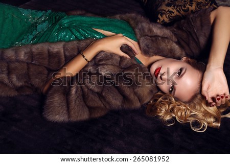 fashion photo of gorgeous woman with blond hair  in elegant dress and fur coat lying on bed