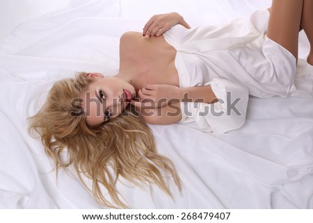 fashion studio photo of sexy beautiful woman with long blond hair in white male shirt lying on bad