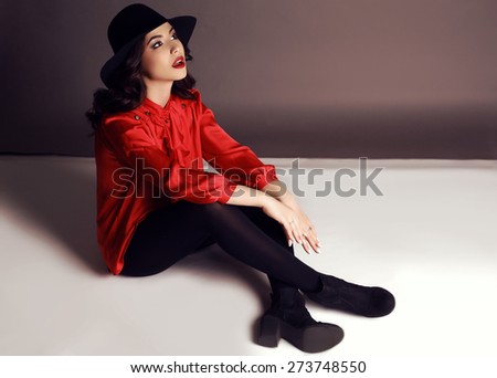 fashion photo of beautiful girl with dark hair in elegant red blouse and black hat posing in studio