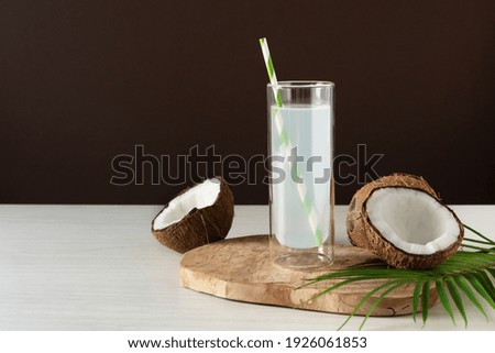 Coconut water in a glass with straw on white wooden table over brown background. Fresh detox coconut juice.
