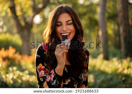 Gorgeous woman eating bar of dark chocolate in the sunny park. Woman enjoying chocolate with her eyes closed 