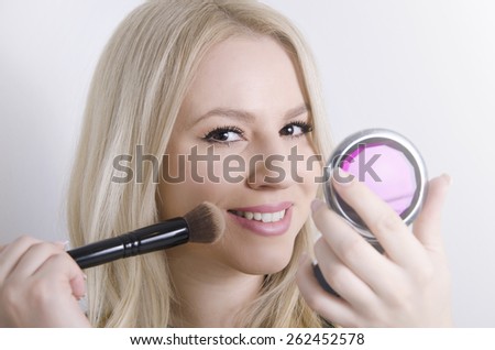 Perfect blonde Caucasian girl putting on makeup, while holding mirror
