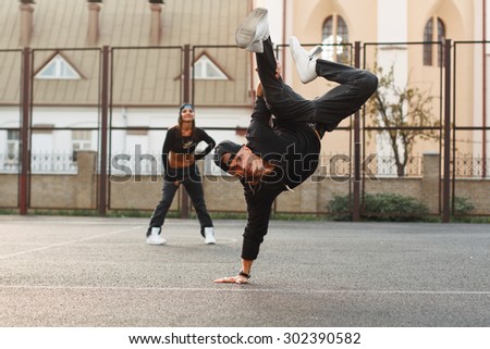 Handsome guy in a stylish black clothes dancing hip-hop. dancer standing on his hand. The girl is behind the guy.