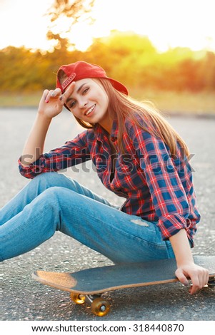 Cheerful girl in a shirt and a baseball cap sitting on the asphalt with a skateboard.Beautiful sunset.