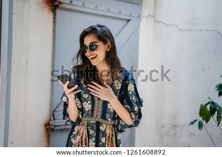 Portrait of a young, attractive and beautiful Indian Asian woman wearing a dress and sunglasses with her smartphone. She looks surprised and delighted and is smiling as she looks at her phone.