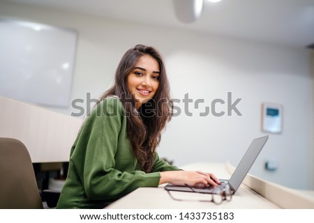Portrait of a beautiful, young and intelligent-looking Indian Asian woman student wearing a white shirt and green tracker smiling as she works on her laptop in a university classroom. 