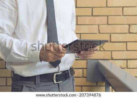 Businessman holding digital tablet computer, standing at the office balcony with yellow brick wall.