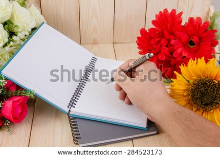 Hand holding a pen with notepad and flower on wood texture.