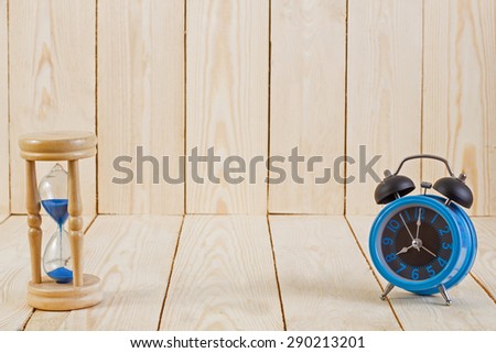 Blue alarm clock and hourglass on wood background.