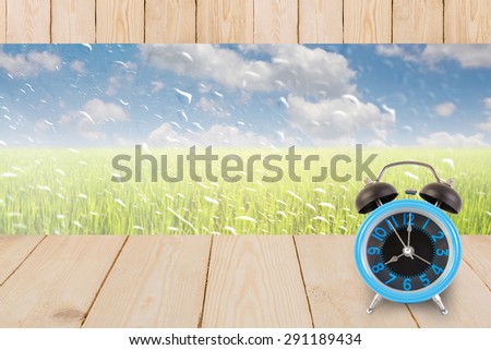 Blue alarm clock on table wood texture with rain drops on a window or water drops on grass blurred with green rice field and blue sky.