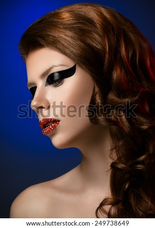 Makeup and beauty theme: portrait of a beautiful young sexy girl with big beautiful eyes, black arrow on the eyes, red lips with diamonds on a dark blue background in studio isolated