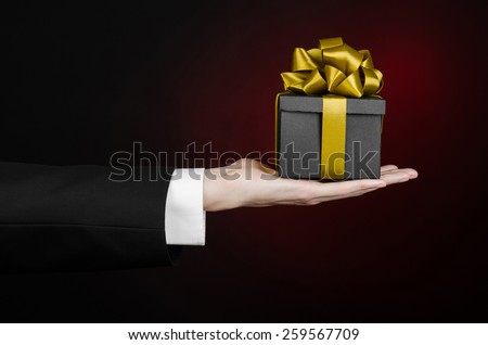 The theme of celebrations and gifts: a man in a black suit holding a exclusive gift packaged in a black box with gold ribbon, beautiful and expensive gift on a dark red background in studio isolated