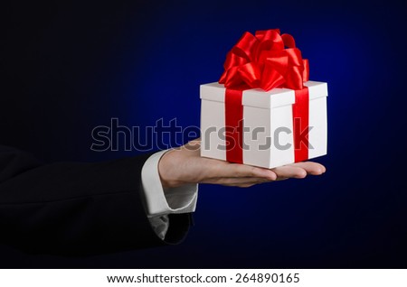 The theme of celebrations and gifts: a man in a black suit holding a exclusive gift wrapped in white box with red ribbon, beautiful and expensive gift on a dark blue background in studio isolated