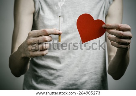 Cigarettes, addiction and public health topic: smoker holds the cigarette in his hand and a red heart on a dark background in the studio