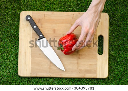 Vegetarians and cooking on the nature of the theme: human hand holding a red pepper and a knife on a cutting board and a background of green grass top view
