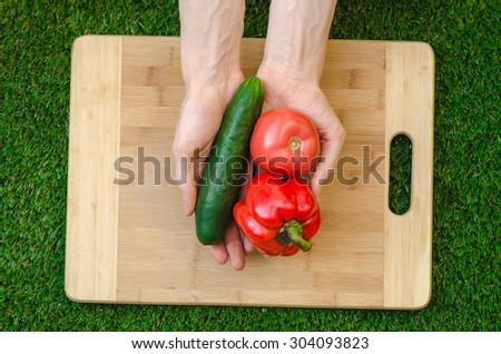 Vegetarians and cooking on the nature of the theme: human hand holding cucumber, tomato and red pepper on a cutting board and a background of green grass top view