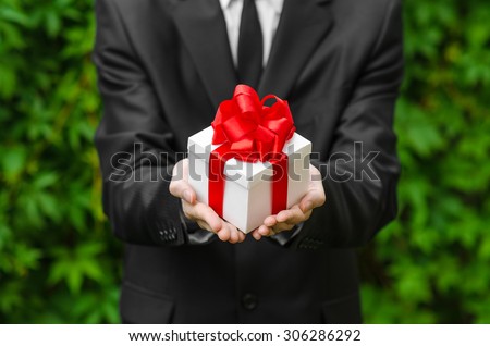 Gift and business theme: a man in a black suit holding a gift in a white box with a red ribbon on a background of green grass