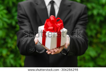 Gift and business theme: a man in a black suit holding a gift in a white box with a red ribbon on a background of green grass