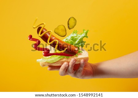 Hand holds hot dog in which sausage, salad, mustard, ketchup, pickle cucumbers are flying on a yellow background