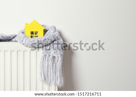 House model wrapped in scarf on radiator indoors, space for text. Winter heating efficiency 商業照片 © 