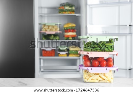 Containers with different frozen vegetables on white marble table near open refrigerator