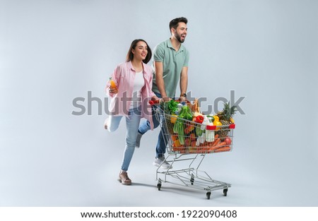 Young couple with shopping cart full of groceries on grey background