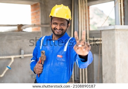 Happy young indian engineer or repairman holding wrench showing victory sign or hand gesture by looking at camera - concept of professional occupation,Confidence and blue collar job. 商業照片 © 