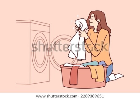 Housewife woman sits near washing machine and inhales fragrant smell freshly washed towel after using good laundry detergent. Girl takes out washed clothes from washing machine putting them in bowl 