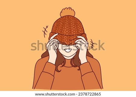 Woman covers face with knitted hat with pompom and smiles standing in warm sweater. Happy girl rejoices at onset of winter or autumn cold weather and puts on handmade hat given by loved one