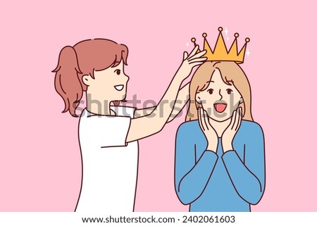 Little girl places crown on overjoyed sisters head while playing princesses from ancient kingdom. Two sisters from junior school try on golden crown received for achievements in education